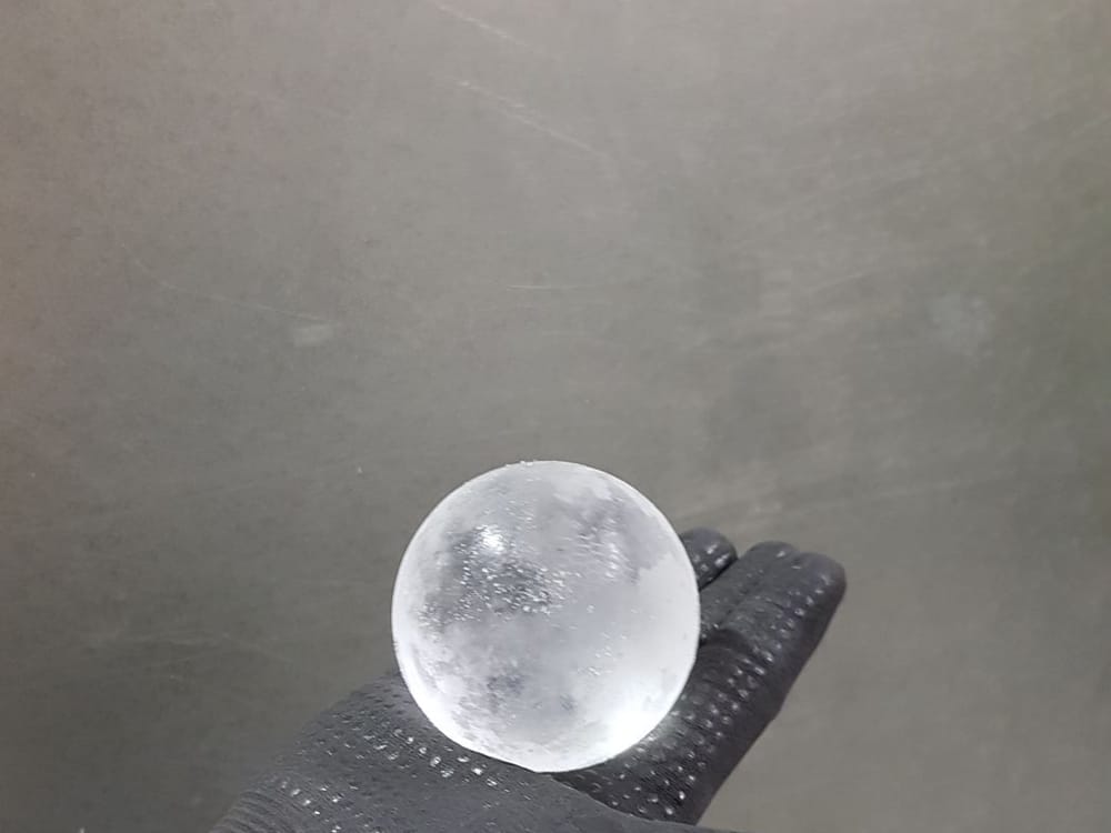 A clear, spherical laboratory hailstone held in a gloved hand before a test shot