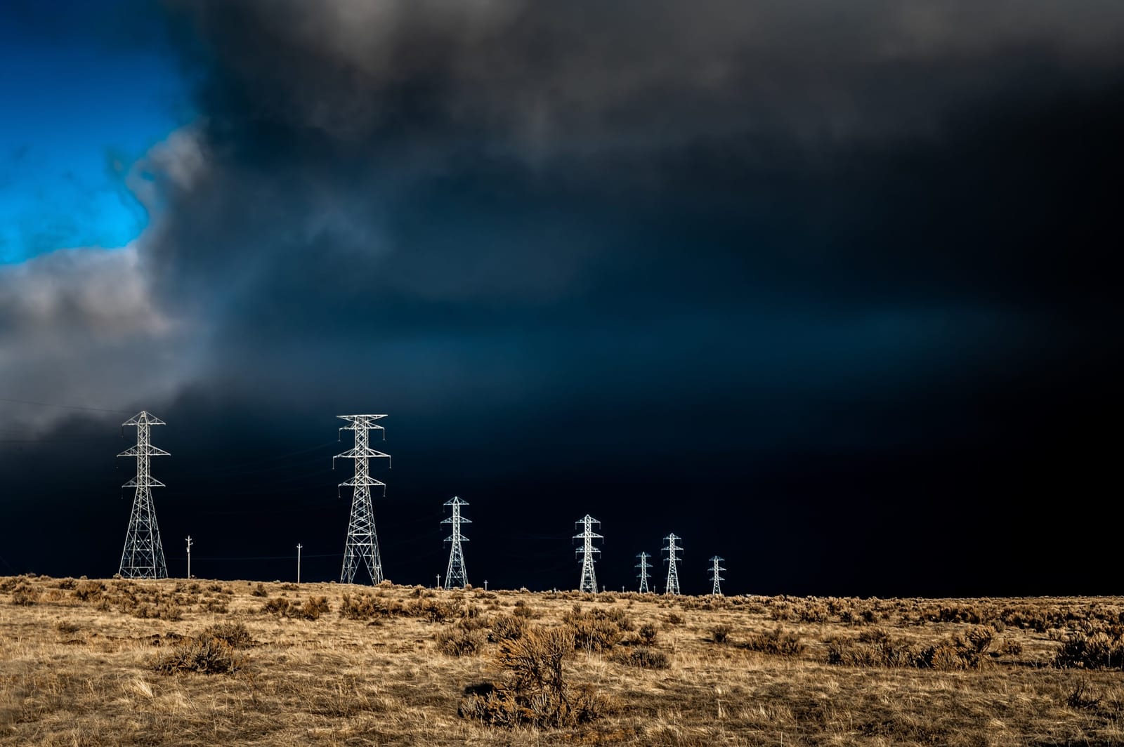 A dark storm front over a line of transmission towers on dry grassland.