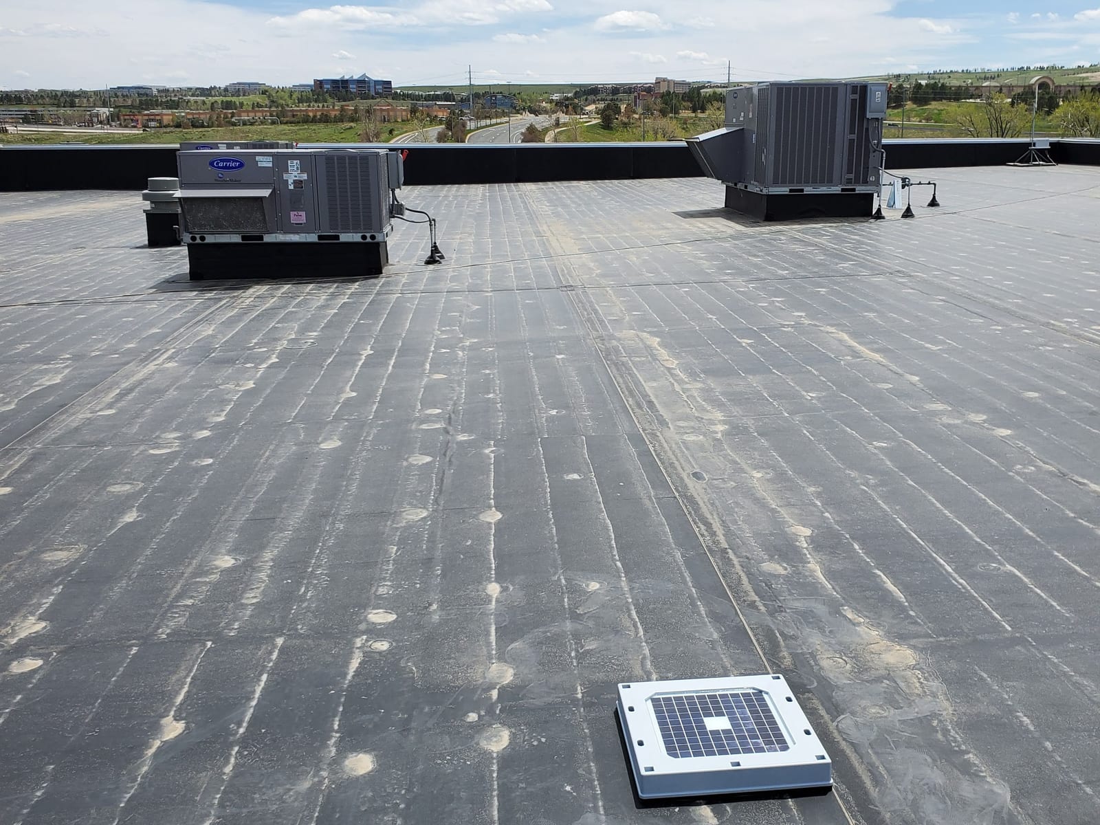 A Hailios Tessera sensor on a flat commercial membrane roof beside rooftop HVAC units, a business park on the horizon