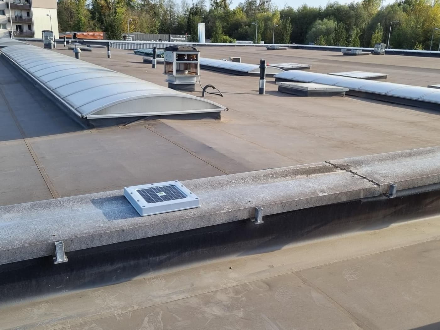 A Hailios Tessera sensor on the parapet of a large flat commercial roof, rows of barrel skylights and rooftop vents behind it