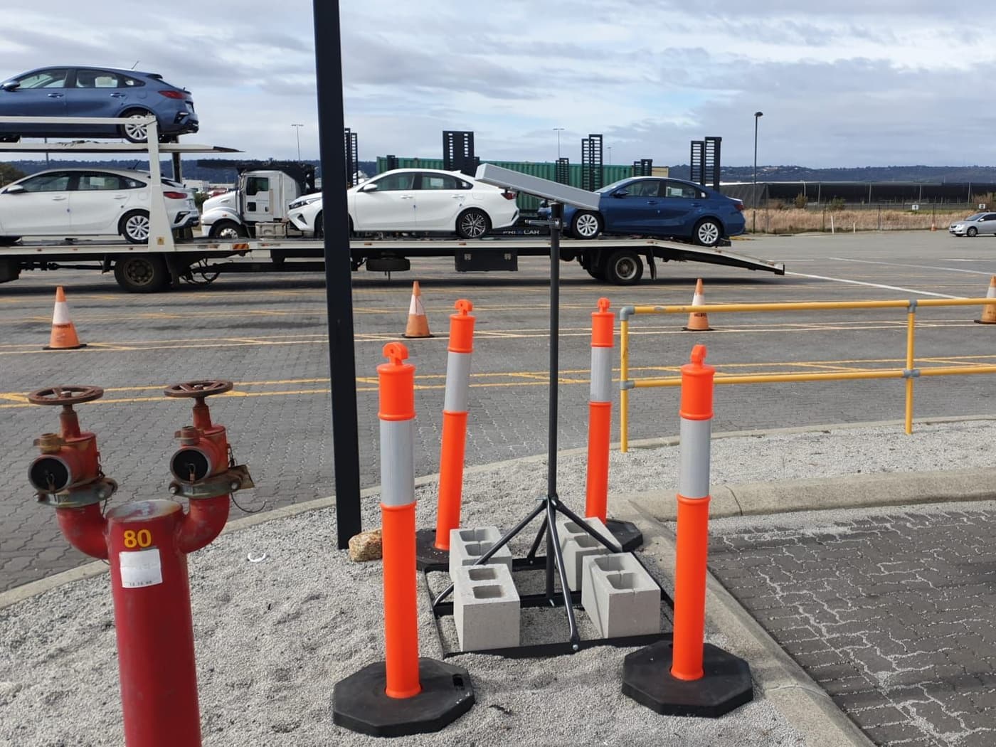 A Tessera sensor on a weighted stand at a vehicle staging yard, a car carrier loaded with new cars behind it
